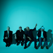 Members of the band the pixies sitting on the floor of a studio with blue lighting 